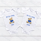 Two white baby rompers with teddy bear graphics and text on a light wooden surface.