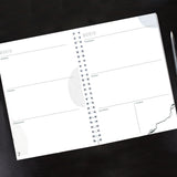 Spiral-bound planner on a black surface with a pen to the right.