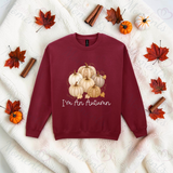 A maroon crewneck sweatshirt with a graphic of pumpkins and the text 'I'm An Autumn' in white script, laid flat on a white textured blanket with autumn props like leaves, cinnamon sticks, and small pumpkins.