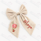 Beige bow with a teddy bear and hot air balloon design and the name 'Maya' on a white background.