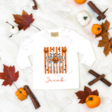 A white long-sleeve baby t-shirt with a Halloween graphic of skeletons behind orange pumpkins and the name 'Jacob' printed below, styled in a flat lay with autumn leaves, pumpkins, cinnamon sticks, and white pumpkins on a white fabric background.
