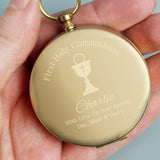 Gold pocket watch engraved with 'First Holy Communion Charlie' held in a hand against a gray background