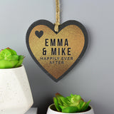 Heart-shaped slate plaque with 'EMMA & MIKE HAPPILY EVER AFTER' text, hanging against a grey background with potted plants.