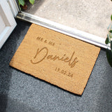 A personalised rectangular indoor doormat with a brown coir surface and personalisation. The doormat is placed on a wooden floor and has a plant in the background. Personalisation reads "Mr & Mr Daniels 19.02.24"