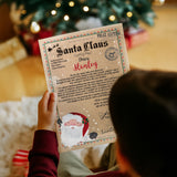 Image of child holding and reading their A4 Personalised letter from Santa Claus. Features the name "Stanley" on the letter and blurred, out of focus Christmas Tree in background.