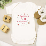 Baby onesie with 'Amelia's First Christmas 2023' text on a light surface with wooden blocks and baby booties.