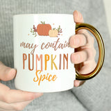 Female hands holding a white mug with gold handle, featuring the phrase "may contain pumpkin spice"