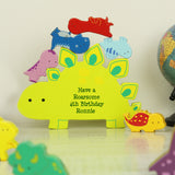 Colorful dinosaur-shaped wooden toy with 'Have a Roarsome 4th Birthday Ronnie' text, surrounded by small dinosaur toys on a light surface.