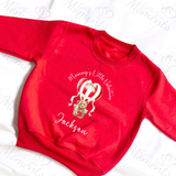 Red Toddler Crewneck sweatshirt with image of a teddy bear in a heart shaped hot air balloon with banner through it saying "Happy Valentines Day". Above the image, curved around it, are the words "Mummy's Little Valentine" in white writing and the name Jackson appears beneath. Sweatshirt is laid flat on a white background with arms out to the side.