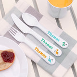 Personalized children's cutlery set with names and dinosaur illustrations on a table with a plate and mug.