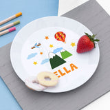 White plate with colourful mountain and hot air balloon design, topped with a strawberry and biscuits, on a grey surface.