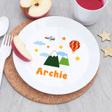 Plate with 'Archie' and mountain design, surrounded by apples and a cup on a grey surface.