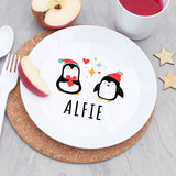 Round white plate with cartoon penguins and 'ALFIE' text on a cork placemat, surrounded by apples and a cup.