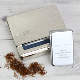 A silver tobacco tin and a silver lighter with personalized engravings, displayed alongside some tobacco leaves.