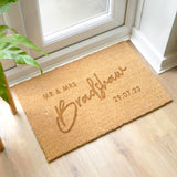 A personalised rectangular indoor doormat with a brown coir surface and personalisation. The doormat is placed on a wooden floor and has a plant in the background.
