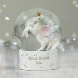 A clear glass snow globe with a white unicorn figurine inside, placed on a white base that has 'Shine Bright Isla' written on it.