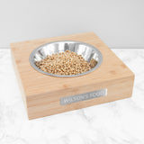 A personalised bamboo pet bowl with a metal tag, containing some food. The bowl is placed on a marble surface, and has engraved text of the pets name, reading "Wilson's Food"