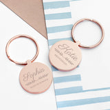 Two personalised rose gold keyrings with engraved names 'Sophia' and 'Katie' on a paper with a striped background. Beneath the name Katie is the role "Bridesmaid" and beneath Sophia is the role "Maid of Honour"