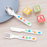 A set of personalised kids' cutlery with colorful shapes on the handles, including a spoon, fork, and knife, presented on a wooden table with a blue plate and toy blocks in the background.