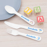 A set of children's cutlery with a pastel blue and yellow color scheme, including a fork, knife, and spoon, each with 'Annabelle' and various playful designs such as a rainbow and clouds.