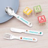 A personalised kids' cutlery set laid out on a wooden table, consisting of a fork, knife, and spoon, with each piece having a colorful character or object design and the name 'Charlotte' printed on them.