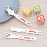 A set of children's cutlery with a spring bunny theme, including a fork, knife, and spoon, laid out on a table with a blue plate and colorful wooden blocks in the background.