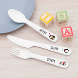 A set of personalized kids' winter penguin cutlery, including a fork, knife, and spoon, with penguin designs on each piece, placed on a wooden surface next to a blue plate and a toy wooden blocks.